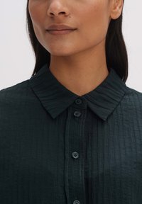 Dark green, textured shirt featuring a collared neckline and front button closure, with subtle vertical stripes and black buttons.