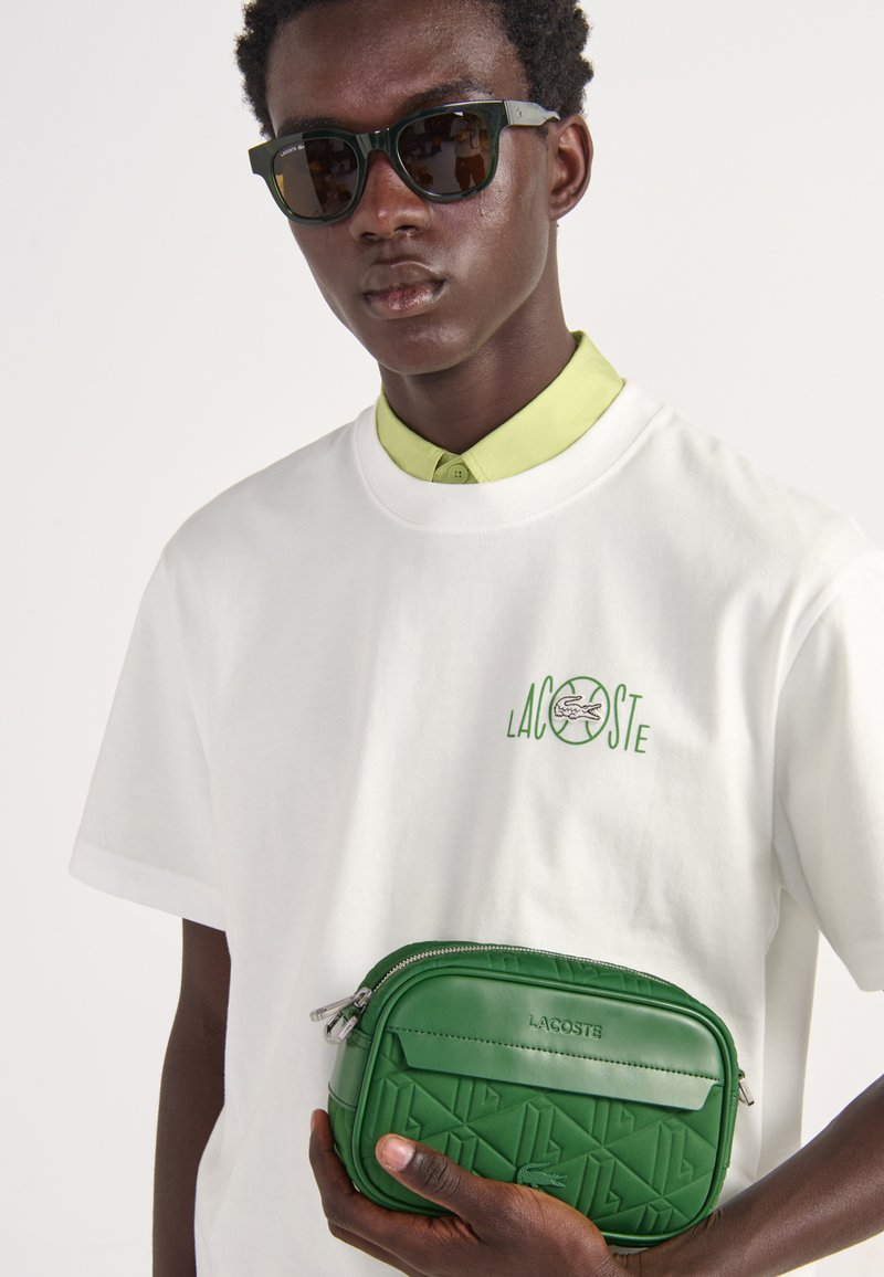 Jeune homme portant des lunettes de soleil noires et un t-shirt blanc Lacoste, tenant un sac bandoulière matelassé vert Lacoste sur un fond uni.