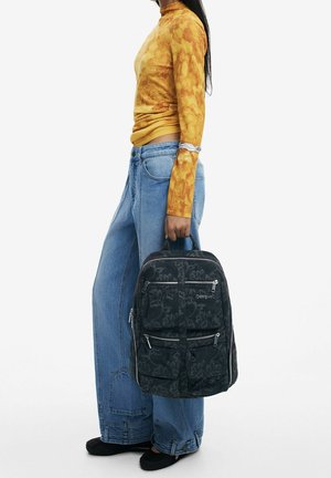 Czarny plecak z tkaniny z kwiatowym wzorem, wieloma kieszeniami na zamek błyskawiczny i górnymi uchwytami. Noszony z luźnymi dżinsami z denimu i żółtą bluzką z długim rękawem.