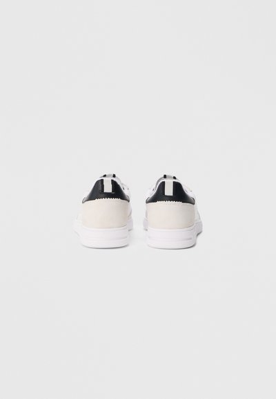 Par de ténis brancos e bege vistos por trás, com detalhes pretos no calcanhar e alças verticais numa superfície simples de cor clara.