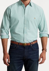 Uomo con camicia azzurro chiaro a maniche lunghe arrotolate, jeans blu scuro, cintura marrone con fibbia dorata e orologio in pelle marrone.