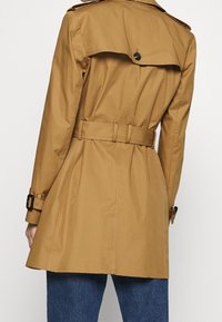 Brązowy płaszcz trench z paskiem w talii, dużym kołnierzem i detalami guzika na plecach. Wykonany z gładkiego, wodoodpornego materiału.