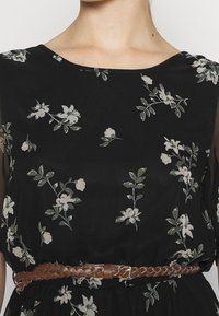 Musta kukkamekko, jossa on vaaleanpunaisia ja vihreitä kukkakuoseja, sileä kangas ja punottu ruskea vyö vyötäröllä.