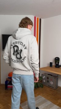Moleton cinza com uma impressão gráfica preta nas costas, combinado com jeans azul claro; sofá, plantas e uma mesa pequena visíveis ao fundo.