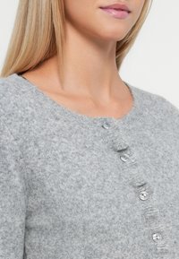 Hallide hall cardigan, mille ringikrae, seitsme ümmarguse nööbiga ja pehme, tekstuuriga viimistlusega, kus on kerge volang nööpide real.