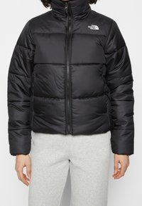 Chaqueta negra acolchada con cuello alto, cierre de cremallera y secciones acolchadas. Tejido suave y liso con un detalle de logo en el lado izquierdo del pecho.