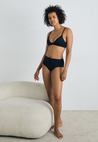 Ensemble de lingerie noire comprenant un bralette triangulaire et des culottes taille haute, avec un tissu lisse et un design minimaliste. Le modèle se tient sur une surface texturée.