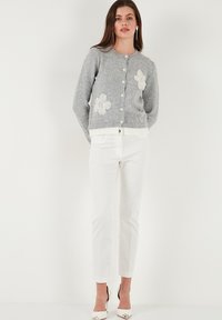 Cardigan gri tricotat cu broderie florală albă, decolteu rotund, închidere cu nasturi și manșete canal. Purtați-l cu pantaloni albi, croiți.