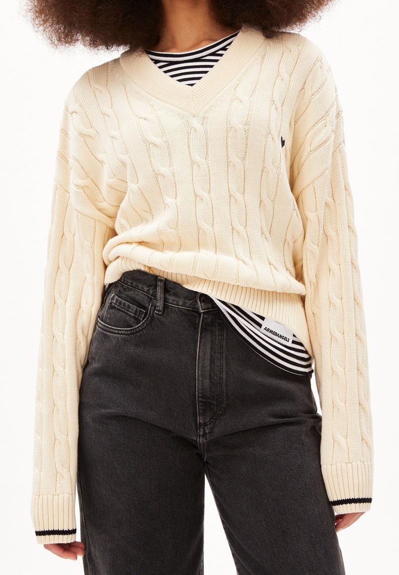 Pull crème en maille torsadée avec un col en V, associé à un jean noir taille haute. En dessous, une chemise rayée noire et blanche est visible.