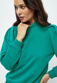 Felpa teal con collo alto, caratterizzata da maniche a sbuffo e polsini a coste. Tessuto liscio, vestibilità comoda, adatta per il tempo libero.