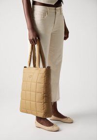 Padded tote bag in tan, featuring a quilted design with vertical lines and two sturdy handles. Worn with light-colored pants and flats.