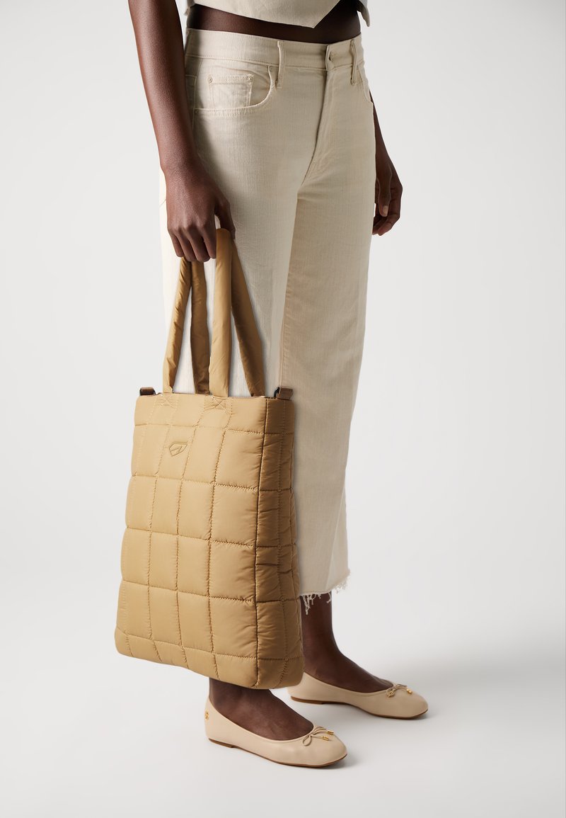 Padded tote bag in tan, featuring a quilted design with vertical lines and two sturdy handles. Worn with light-colored pants and flats.