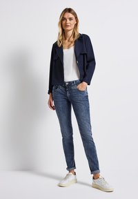 Chaqueta corta azul marino con solapas anchas, combinada con una camiseta sin mangas blanca y jeans azules oscuros. Presenta puños remangados y zapatillas blancas.