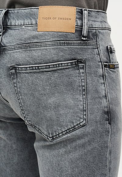 Κοντινό πλάνο πίσω τσέπης γκρι τζιν με δερμάτινη ετικέτα στη μέση που γράφει "Tiger of Sweden".