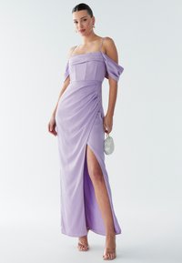 Femme portant une robe lilas à épaules dénudées avec une fente sur la cuisse, des talons à lanières transparentes, des boucles d'oreilles créoles en or, et tenant un petit sac à main blanc.