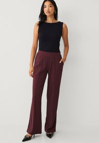 Top negro sin mangas combinado con pantalones anchos color granate con bolsillos laterales. La modelo lleva tacones negros de punta afilada. Textura de tela suave.