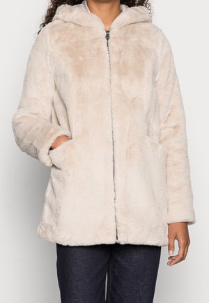 Vrouw draagt een licht beige pluche jas met rits en capuchon, met handen in de zakken en een donkere broek, tegen een effen achtergrond.