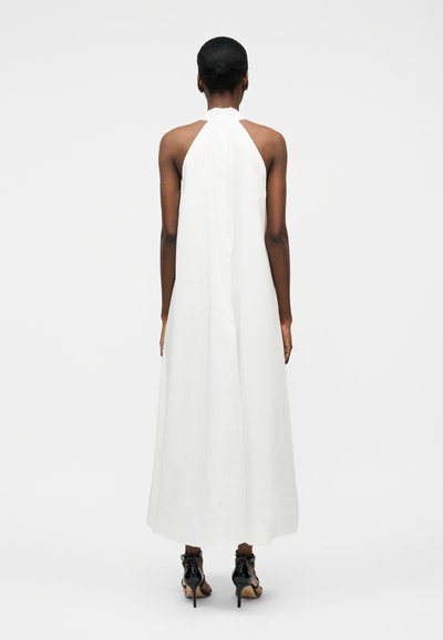 Femme à la peau foncée et aux cheveux attachés, portant une robe blanche sans manches avec un col halter et des sandales à talons hauts noires, debout de dos.