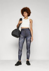 Zwarte draagtas, witte crop top en blauwe leggings met een patroon en een textuur. Model staat rechtop tegen een neutrale achtergrond.