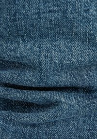 Denimstoff i mellomblått med en teksturert overflate. Har synlige sømmer og svake rynker, noe som skaper et lagdelt utseende.