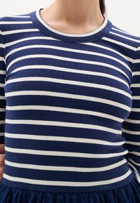 Haut à manches longues rayé bleu et blanc, texture côtelée, encolure ronde, coupe ajustée, associé à un tissu bleu marine froncé à la taille.