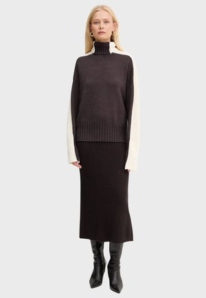 Donna che indossa un maglione a collo alto a maniche lunghe bianco e nero, una gonna nera a coste coordinata e stivali neri fino al ginocchio, in piedi con le braccia rilassate.