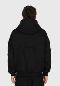 Mann mit lockigem Haar, der von hinten in einem schwarzen Kapuzenpullover vor einem schlichten hellen Hintergrund zu sehen ist.