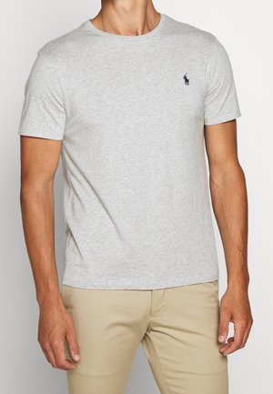 Hombre con camiseta gris claro de manga corta y cuello redondo con pequeño logo oscuro en el pecho izquierdo y pantalones beige, de pie frente a un fondo liso.