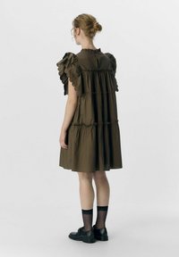 Olivgrünes, gestuftes Kleid mit Rüschensaum und hohem Kragen. Der Stoff ist leicht und glatt. Getragen mit schwarzen Schuhen und durchsichtigen Socken.
