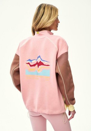 Kobieta o blond włosach, ubrana w różowo-brązową bluzę z blokowaniem kolorów, z wielokolorowym abstrakcyjnym wzorem gór na plecach.