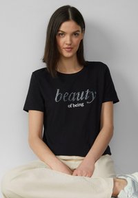 T-shirt noir en coton avec un texte argenté brillant « beauté d'être » sur le devant. Il a un col rond et des manches courtes.