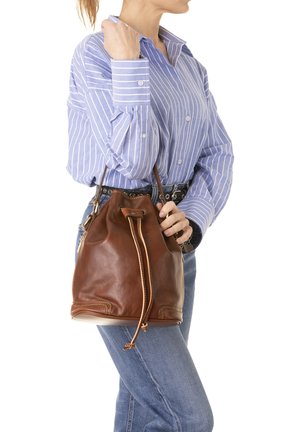Persona con indosso una maglietta a righe blu e jeans tiene una borsa a secchiello in pelle marrone con chiusura a coulisse su sfondo bianco.