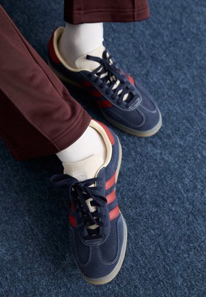 Feet wearing navy and red sneakers with white socks and brown pants on a blue carpeted floor.