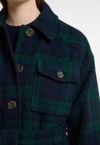 Giacca di lana a quadri blu navy e verde con bottoni, tasche sul petto e cintura in vita. Tessuto testurizzato con un pattern a scacchi ben visibile.