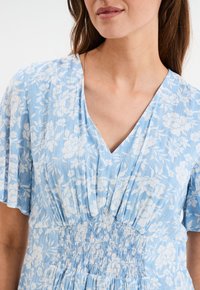 Blaues Blumenkleid mit V-Ausschnitt, gerafftem Taillenbund und kurzen, schwingenden Ärmeln. Der Stoff hat ein strukturiertes, leichtes Design mit weißen Mustern.