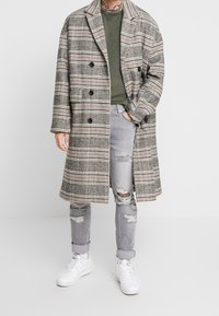 Abrigo largo de cuadros en tonos grises, negros y rojos, con un diseño de doble botonadura, botones negros y grandes bolsillos laterales, sobre una camisa verde y jeans grises desgastados.