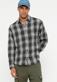 Juoda ir balta languota flanelinė marškiniai su mygtukais, turintis mažą logotipo pleistrą. Modelis dėvi akvamarinės spalvos kepurę ir alyvmedžio žalius cargo džinsus.