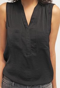 Blouse noire sans manches avec deux poches poitrine et une fente en V, portée avec un jean gris.