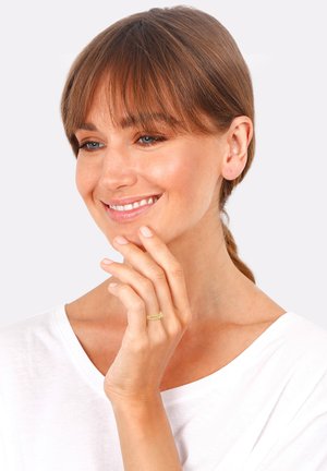 Gold twisted band ring on a model's finger. The model has shoulder-length hair and is wearing a white shirt. Skin tone appears fair.