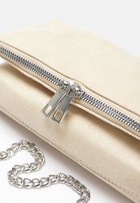 Pochette en daim beige avec double fermeture éclair argentée et bandoulière chaîne argentée sur fond blanc.