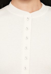 Top bianco a righe con bottoni, caratterizzato da un collo rotondo e sei bottoni abbinati sul davanti, con una texture liscia e morbida.