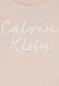 Jasnoróżowa bawełniana bluzka z białym haftowanym napisem "Calvin Klein", miękka w dotyku, z okrągłym dekoltem.