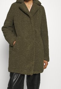 Person klädd i en texturerad olivgrön teddyjacka med krage och fickor, i kombination med svarta läderbyxor mot en slät vit bakgrund.