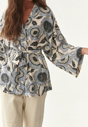 Frau trägt eine beige Bluse mit weiten Ärmeln, schwarz-weißem Blumenmuster und gebundener Taille, kombiniert mit cremefarbenen Hosen.
