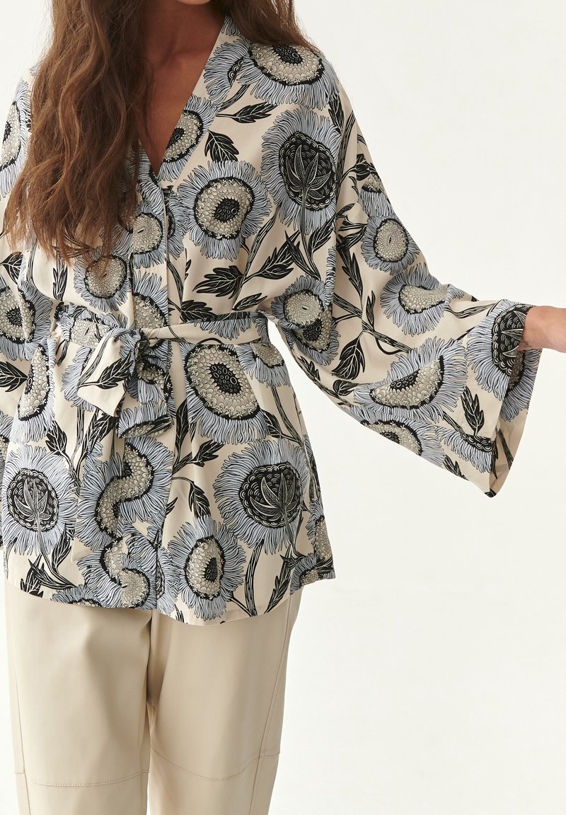 Femme portant un chemisier beige à larges manches avec imprimé floral noir et blanc et taille nouée, associé à un pantalon couleur crème.