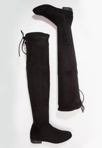 Bottes plates noires au-dessus du genou avec une fermeture éclair sur le côté d'une et un détail de lacet en haut sur fond blanc.