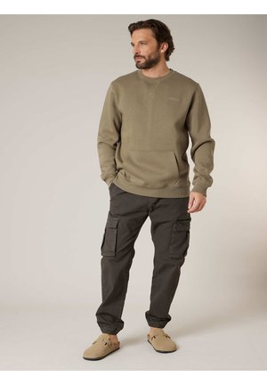 Man draagt een olijfgroene trui, donkere cargobroek en beige instappers, staat met handen in zakken en kijkt naar rechts.