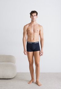 Zwarte boxershorts met een witte tailleband waarop "LACOSTE" staat. De stof lijkt glad en aansluitend, waardoor de lichaamsvorm wordt geaccentueerd.