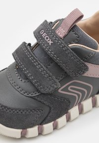 Baskets grises en matière suédée et mesh, avec deux bandes Velcro et une semelle en caoutchouc texturée avec des accents roses. Logo de la marque visible.