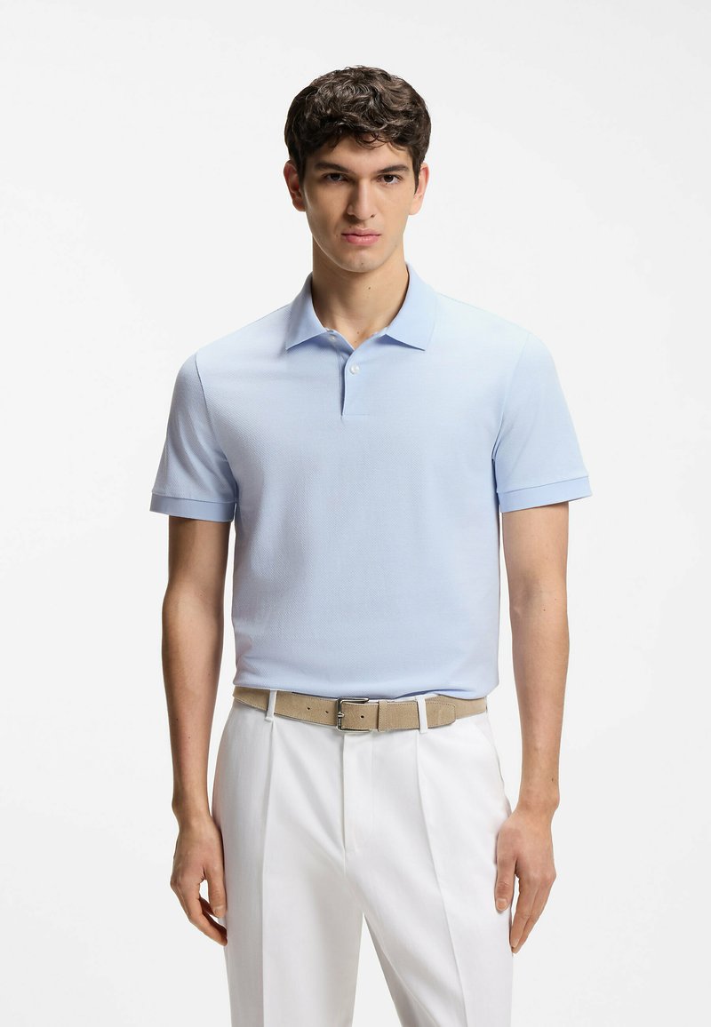 Jeune homme portant un polo à manches courtes bleu clair, ceinture beige et pantalon blanc, debout devant un fond blanc uni.